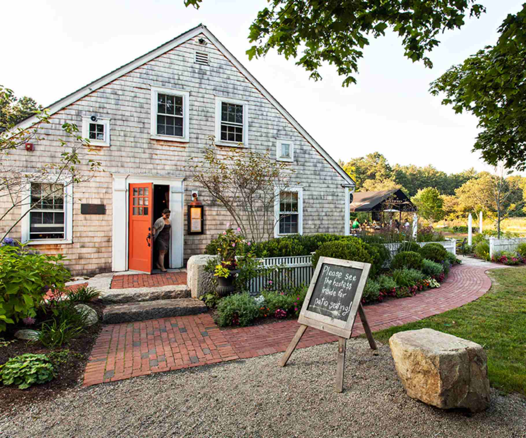 old shingled house with a bright orange door on a brick street with a chalkboard sign in front